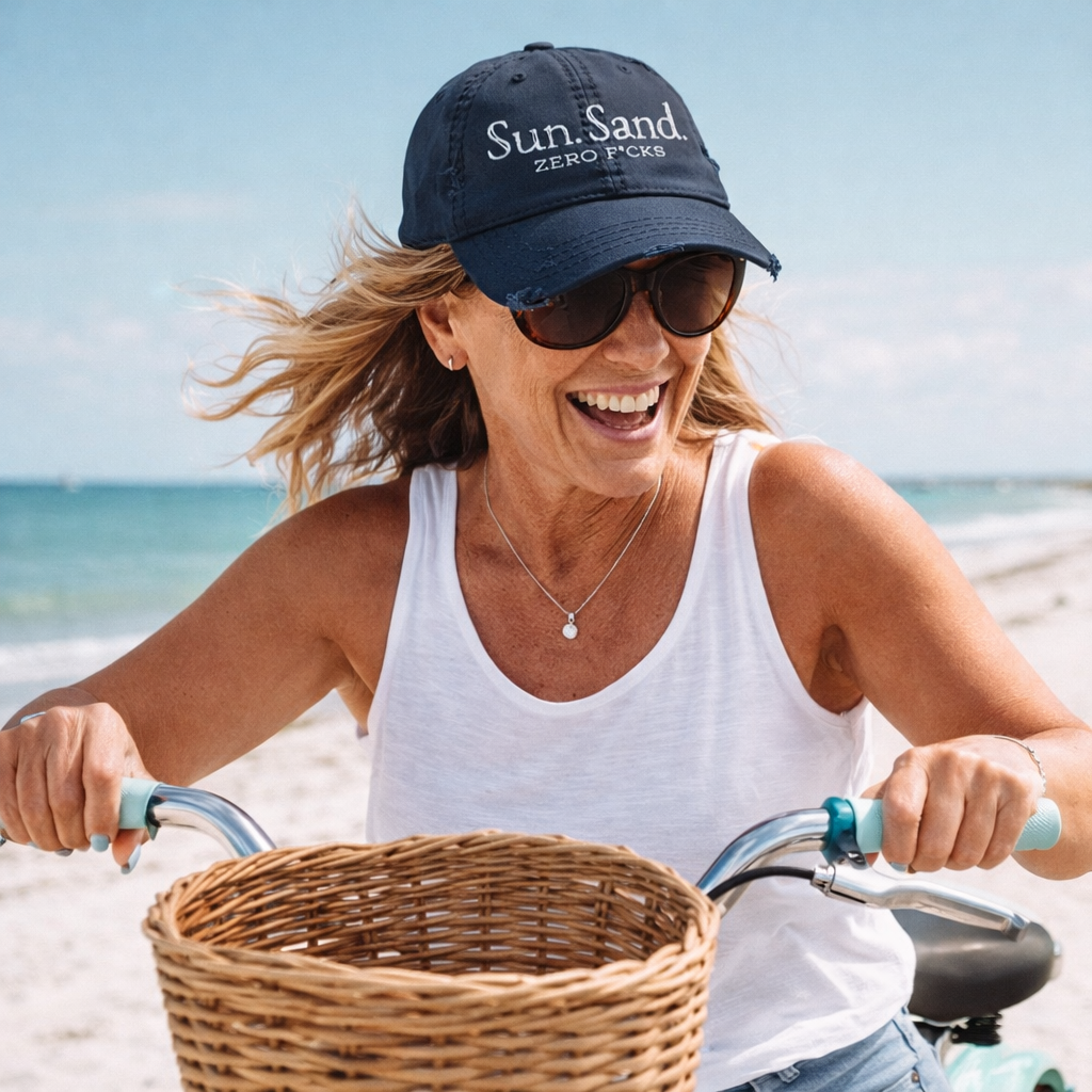 A woman wearing a white tank top and a navy blue cap with the text "Sun. Sand" is riding a bicycle on a beach, smiling and enjoying the sunny day.