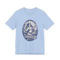 A light blue Maine Salty Girl t-shirt with a graphic design featuring a mermaid or mermaid-like figure surrounded by various elements, including a lighthouse, waves, and a boat.