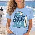 A woman wearing a light blue t-shirt with a graphic design of a surfboard and the text "Surf Bum" printed on it, standing on a beach with the ocean in the background.