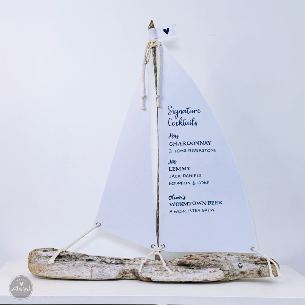 A white sailboat-shaped cocktail menu with a list of signature cocktails, including Chardonnay, Lour Riverstone, Jimmy, Jack Daniels, Bourbon & Coke, and Oliver's Worcester Beer, displayed on a white surface.