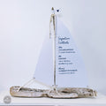 A white sailboat-shaped cocktail menu with a list of signature cocktails, including Chardonnay, Lour Riverstone, Jimmy, Jack Daniels, Bourbon & Coke, and Oliver's Worcester Beer, displayed on a white surface.