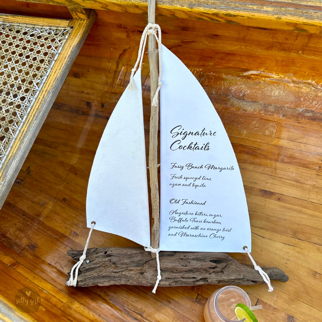 A white sailboat-shaped cocktail menu with a list of signature cocktails, including a "Tropical Cocktail" and "Old Fashioned" drink, placed on a wooden surface.