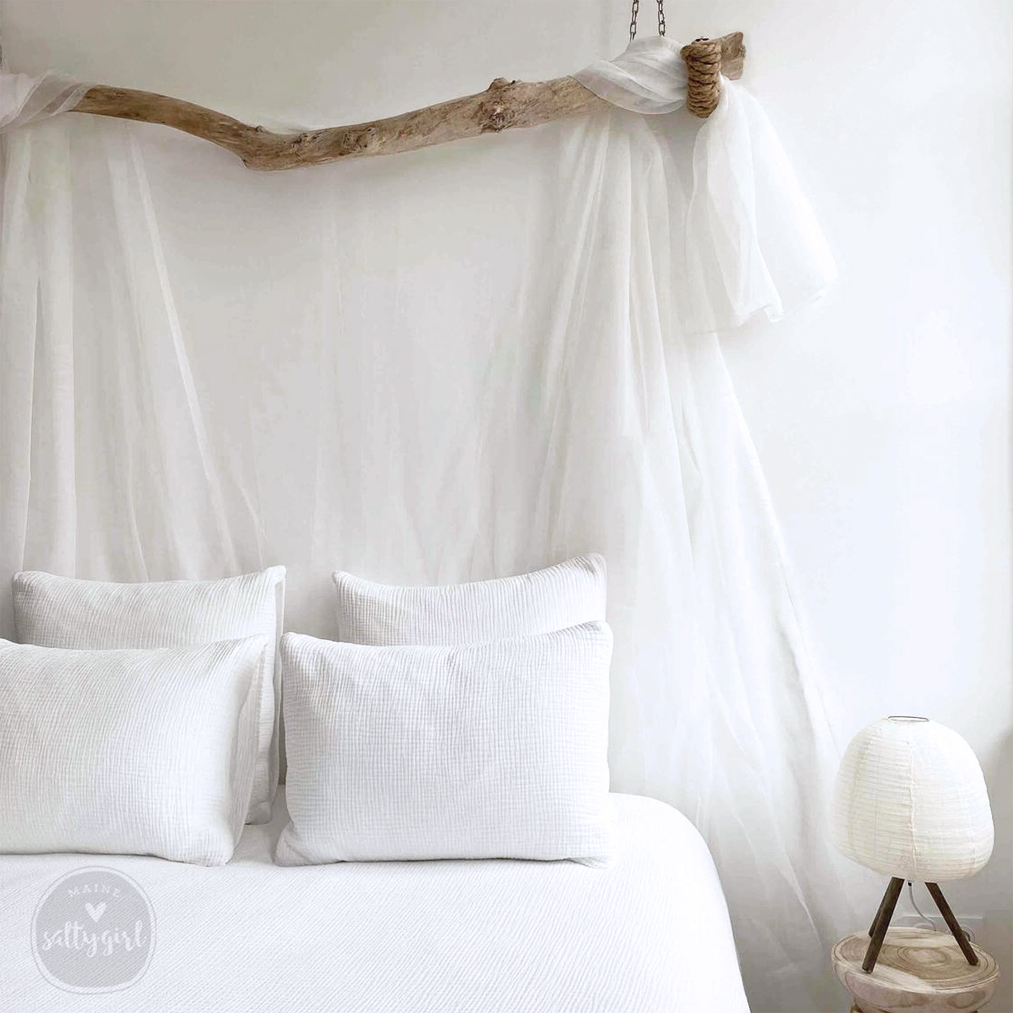 a cozy bedroom with a white bed, a wooden nightstand, and a white lamp. The bed has white pillows and a white canopy, creating a serene and inviting atmosphere.