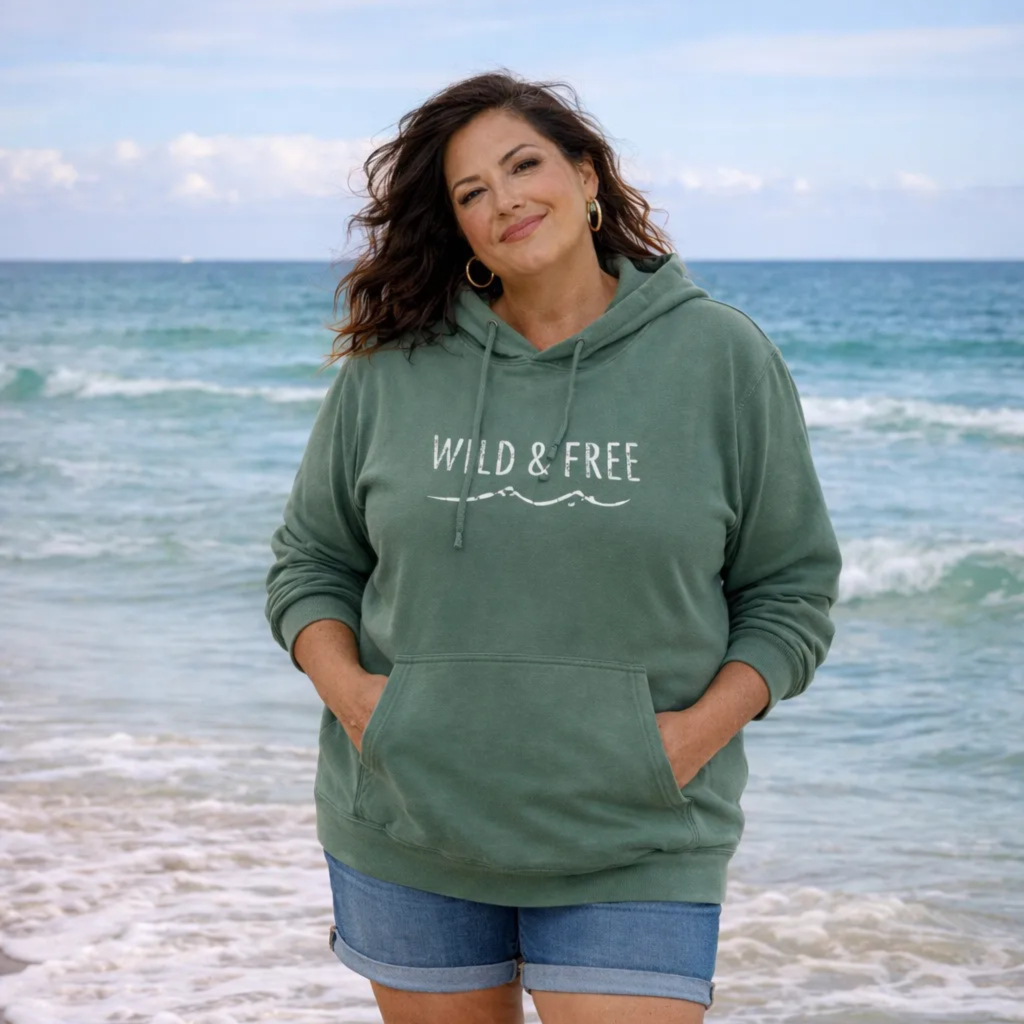 A woman wearing a green hoodie with the text "WILD & FREE" standing on a beach with the ocean in the background.