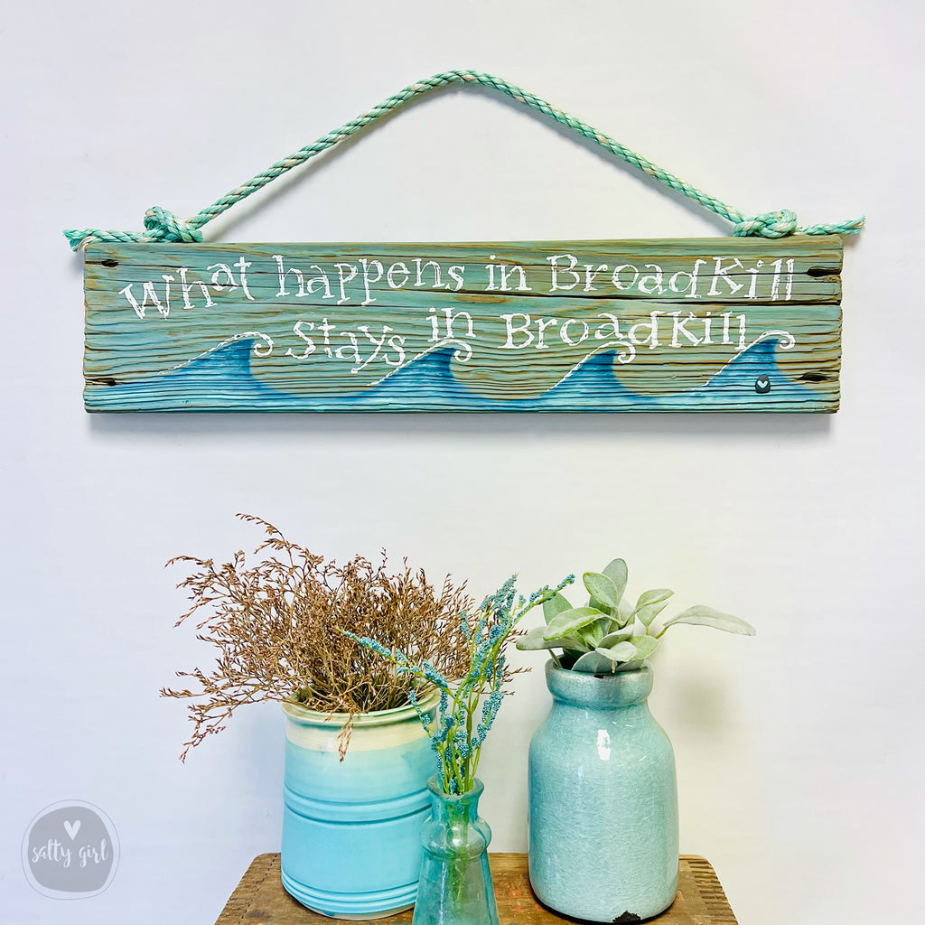 a wooden sign with the phrase "What happens in Broadkills stays in Broadkills" written on it, hanging on a wall. Below the sign, there are two blue vases with dried plants, and a small green plant in a blue vase.