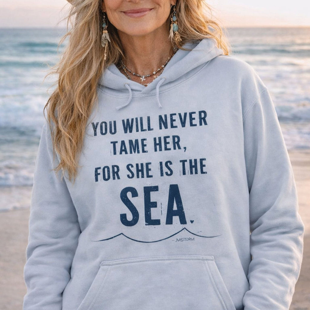 A woman wearing a grey hoodie with a printed message on it, standing on a beach with the ocean in the background.