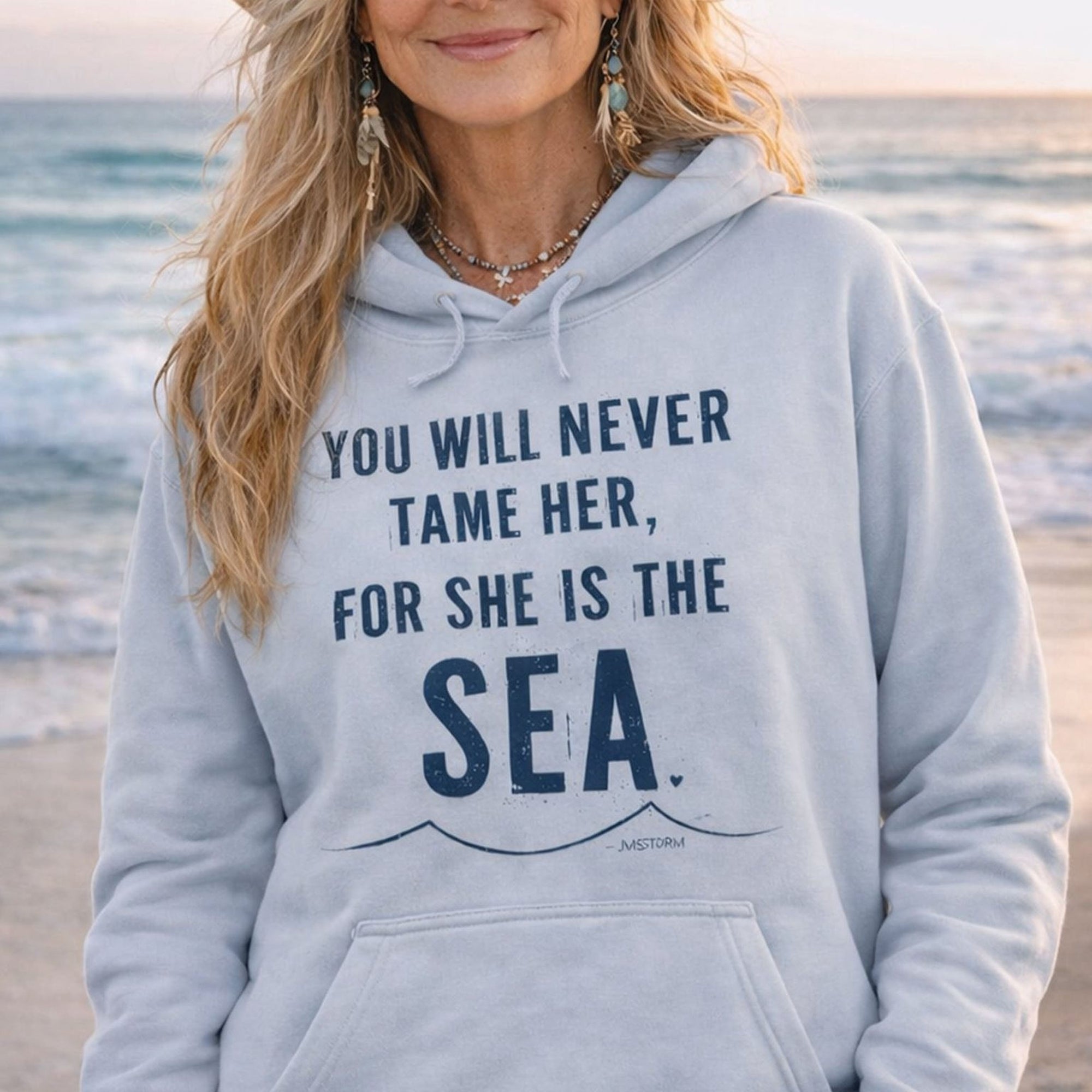 A woman wearing a grey hoodie with a printed message on it, standing on a beach with the ocean in the background.