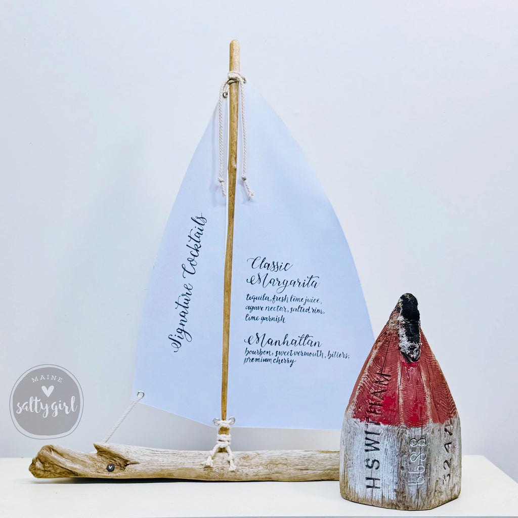 a wooden sailboat-shaped cocktail menu with a red and white striped base, placed on a white surface next to a small wooden object resembling a buoy.