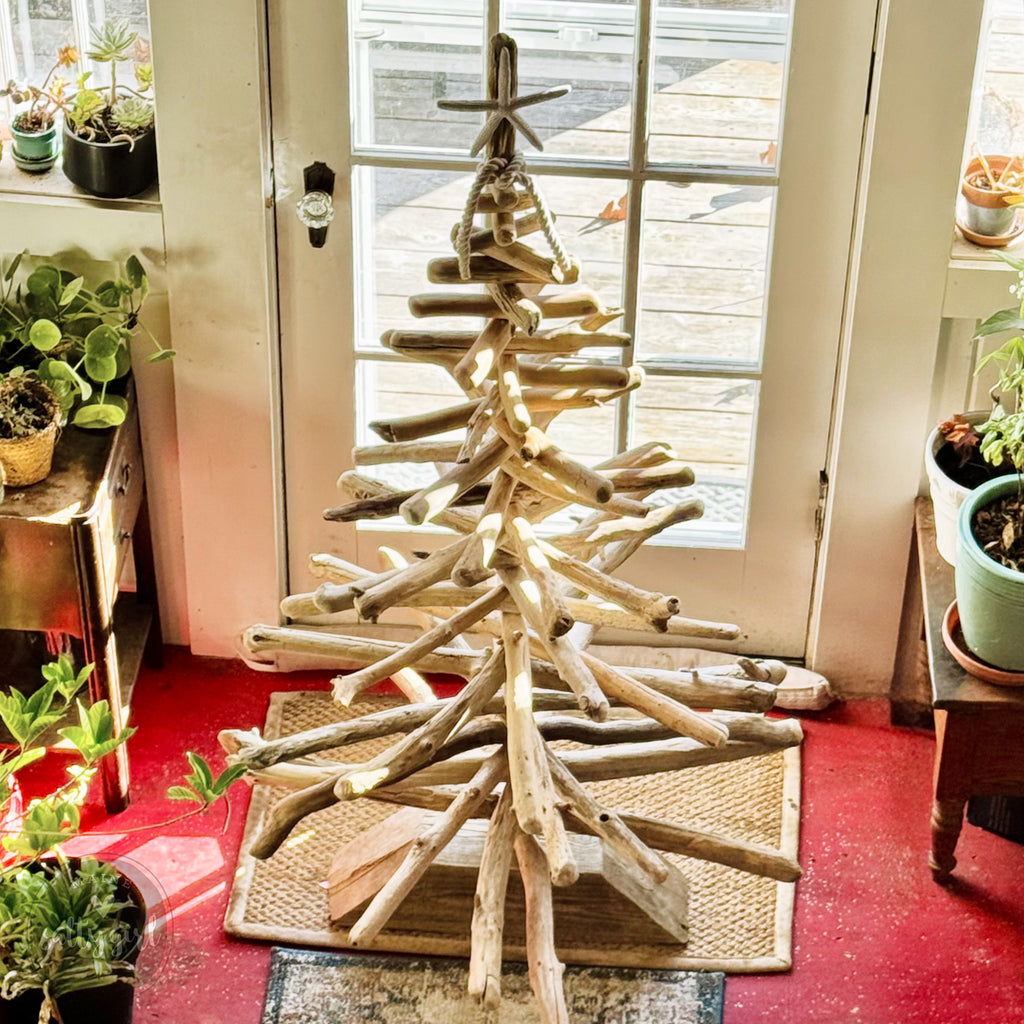 A rustic, handcrafted Christmas tree made from wooden branches and sticks stands in a room with a red floor and a window.