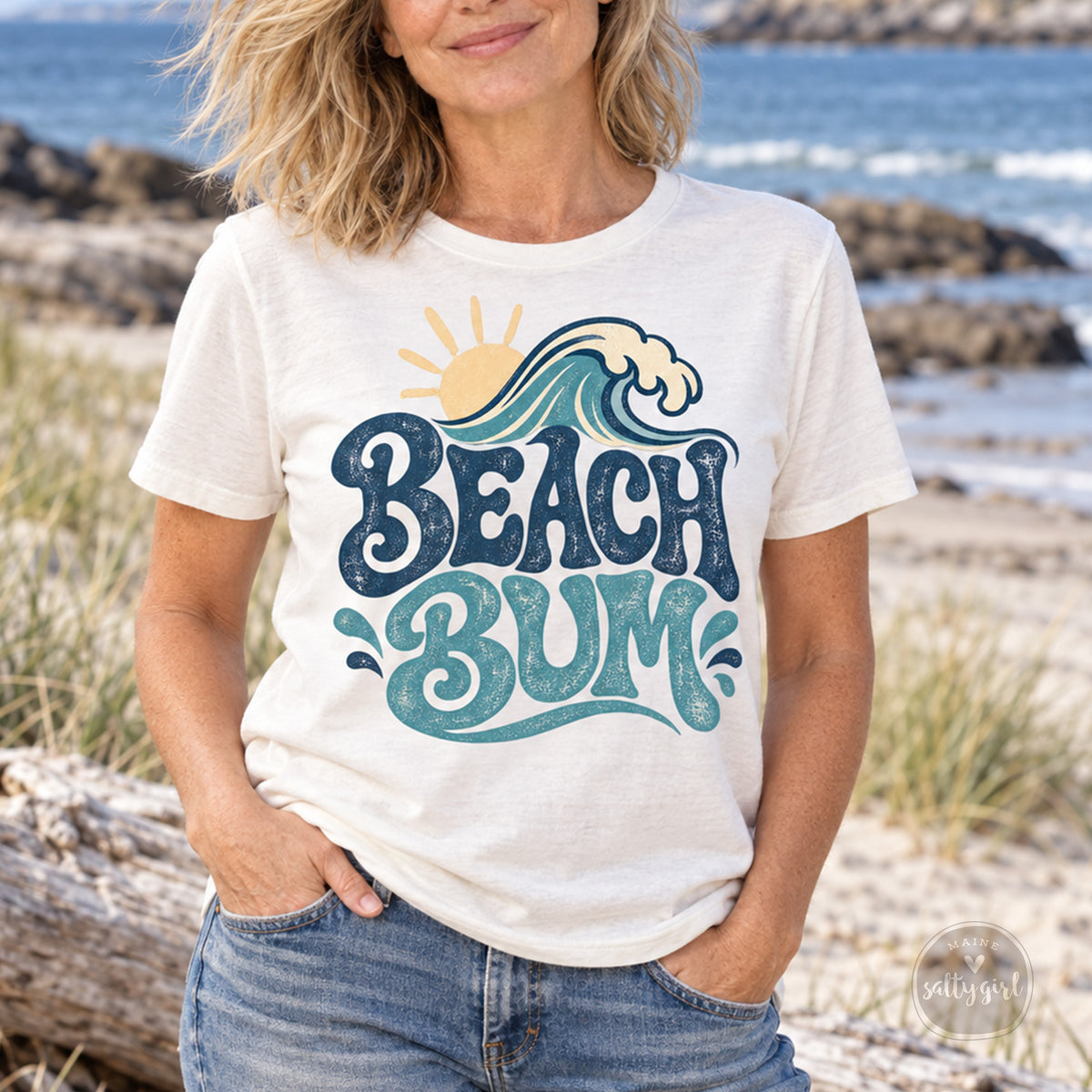 A woman wearing a white t-shirt with a graphic design of a wave and the text "BEACH BUM" printed on it, standing on a beach with the ocean in the background.