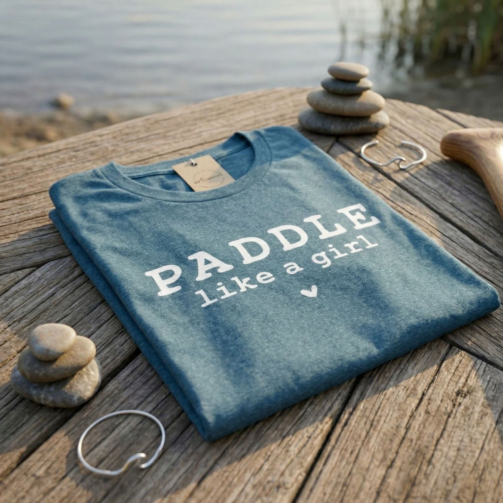 A blue Salty Girl Co t-shirt with the text "PADDLING LIKE A GIRL" printed on it, along with a tag and some small rocks, is placed on a wooden surface.