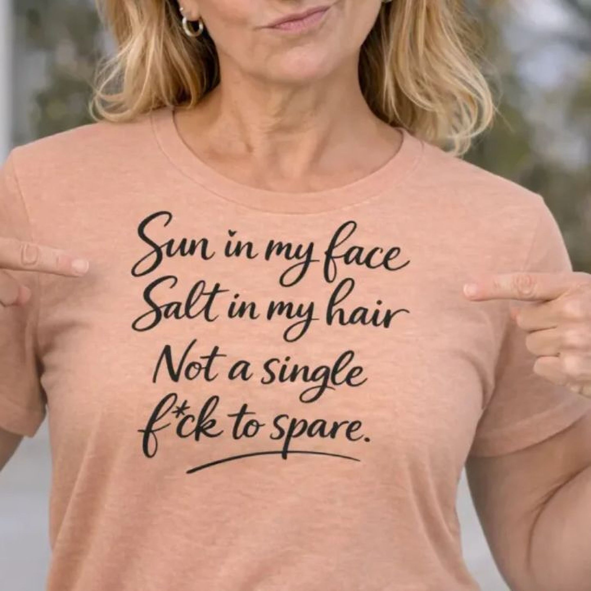 Maine Salty Girl t-shirt with humorous text printed on it saying, sun in my face salt in my hair not a single F * C K to spare, pointing to the text and making a funny facial expression.