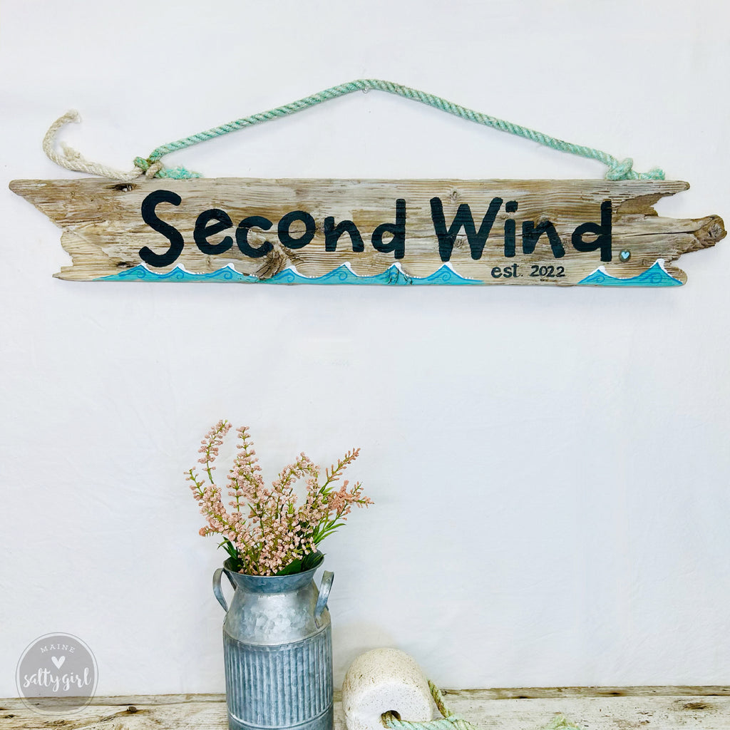 a rustic wooden sign with the name "Second Wind" hanging on a white wall, accompanied by a vase of pink flowers and a small metal can.