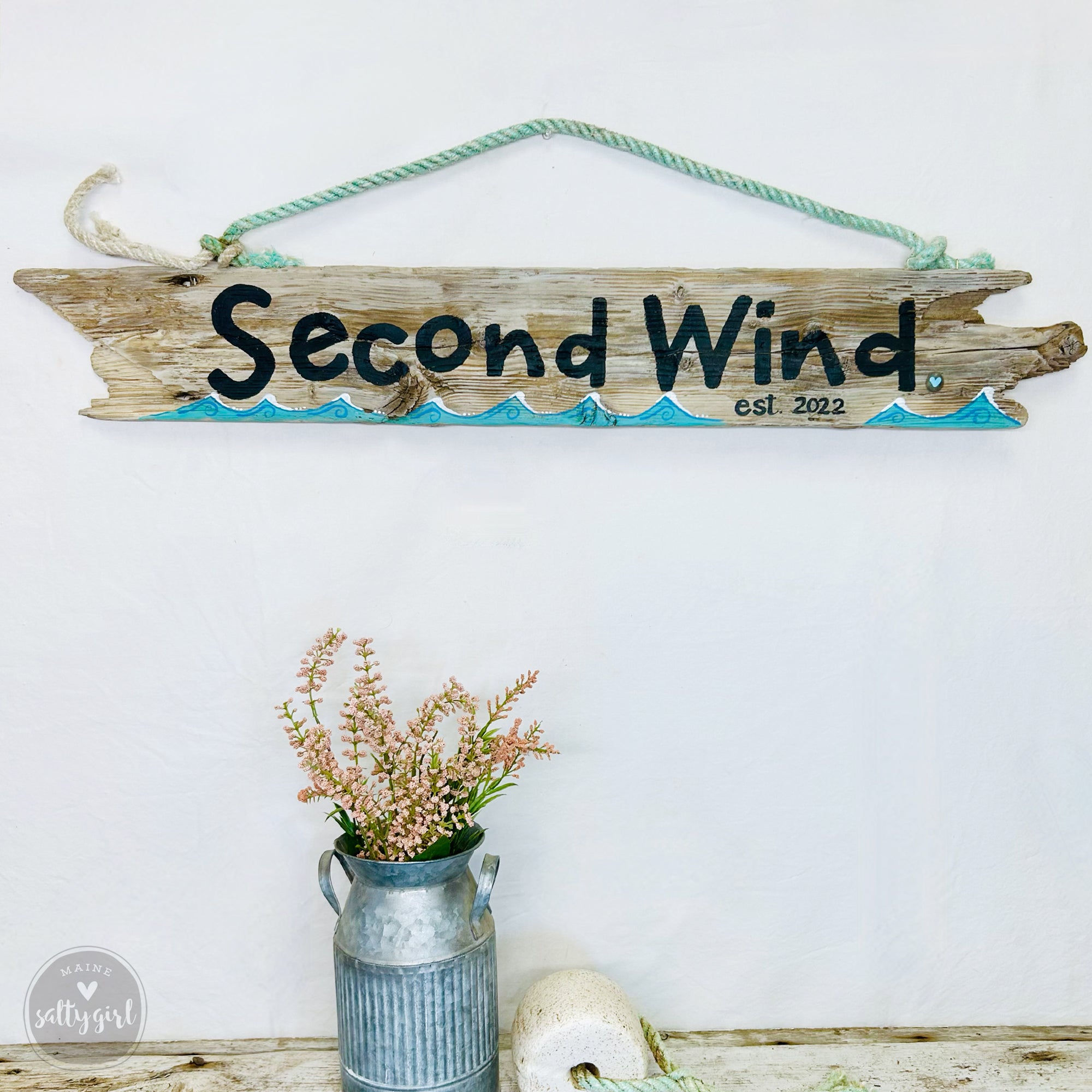 a rustic wooden sign with the name "Second Wind" hanging on a white wall, accompanied by a vase of pink flowers and a small metal can.