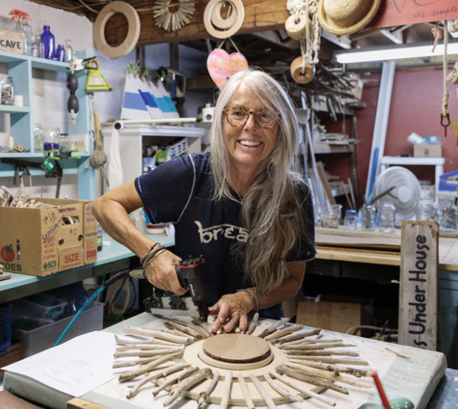 A woman is smiling and working on a craft project in a workshop filled with various tools and materials.