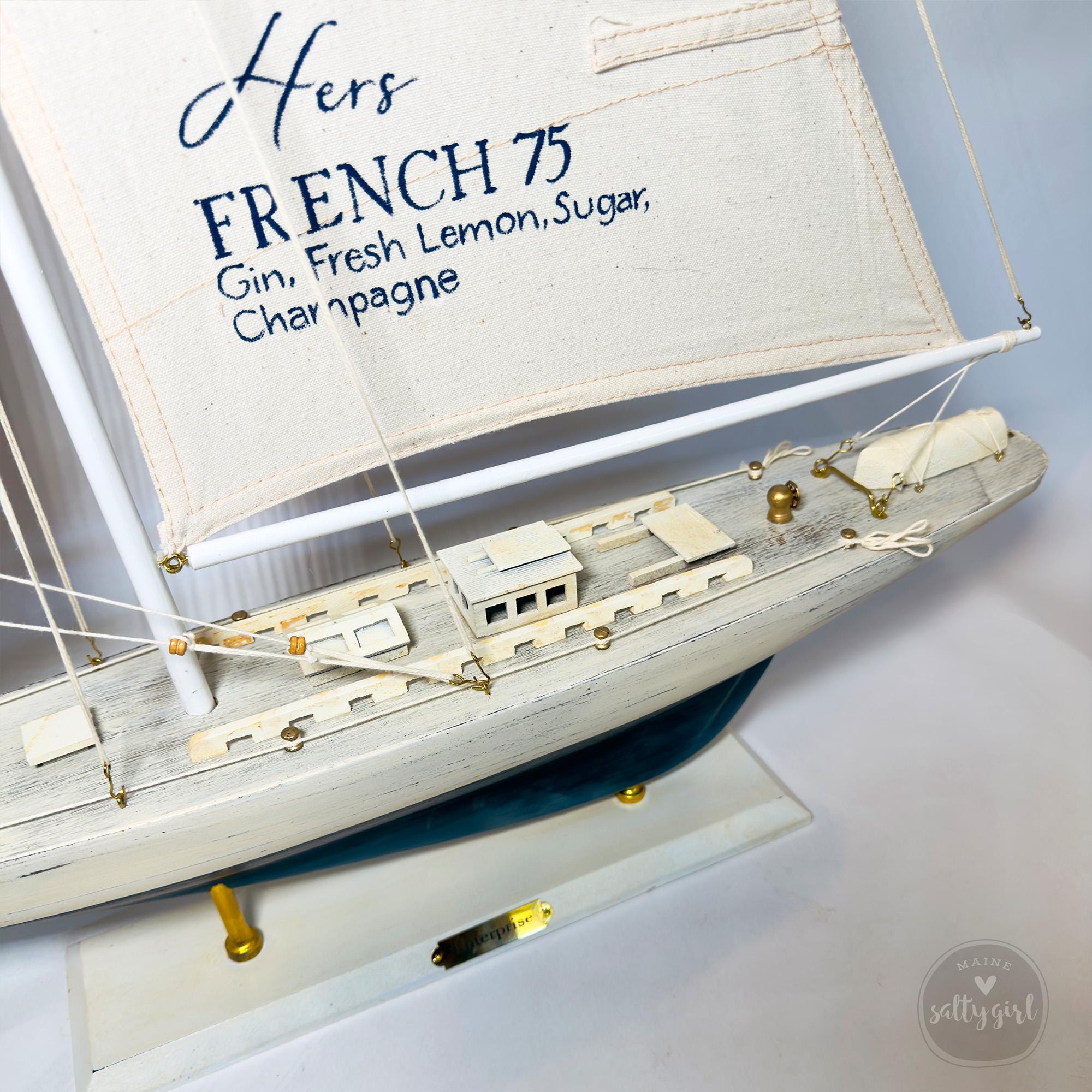 A white model ship with a sail displaying the text "Hers FRENCH 75 Gin, Fresh Lemon, Sugar, Champagne".