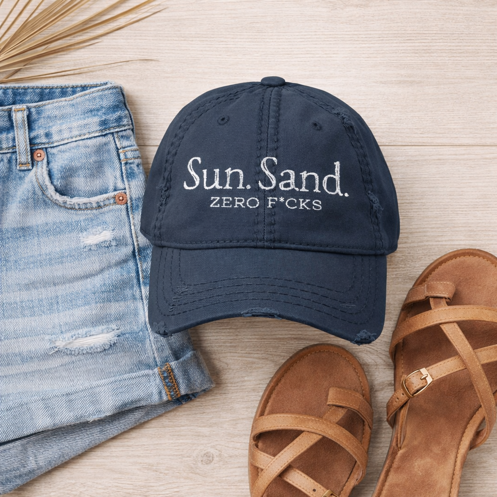 a casual outfit consisting of a blue cap with the text "Sun. Sand. Zero Fucks" printed on it, a pair of blue jeans, and a pair of brown sandals arranged on a wooden surface.