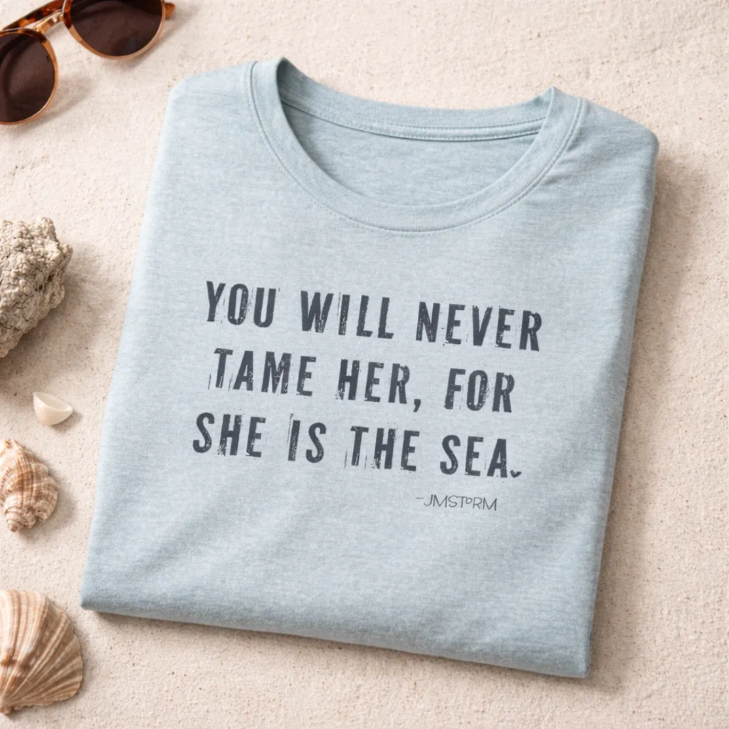 A light blue t-shirt with a quote printed on it, along with some sunglasses and seashells on a sandy surface.