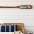 a wooden oar with a boat painted on it, hanging on a white wall above a wicker bench with blue and white striped pillows.
