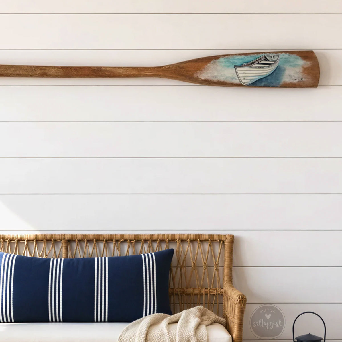 a wooden oar with a boat painted on it, hanging on a white wall above a wicker bench with blue and white striped pillows.