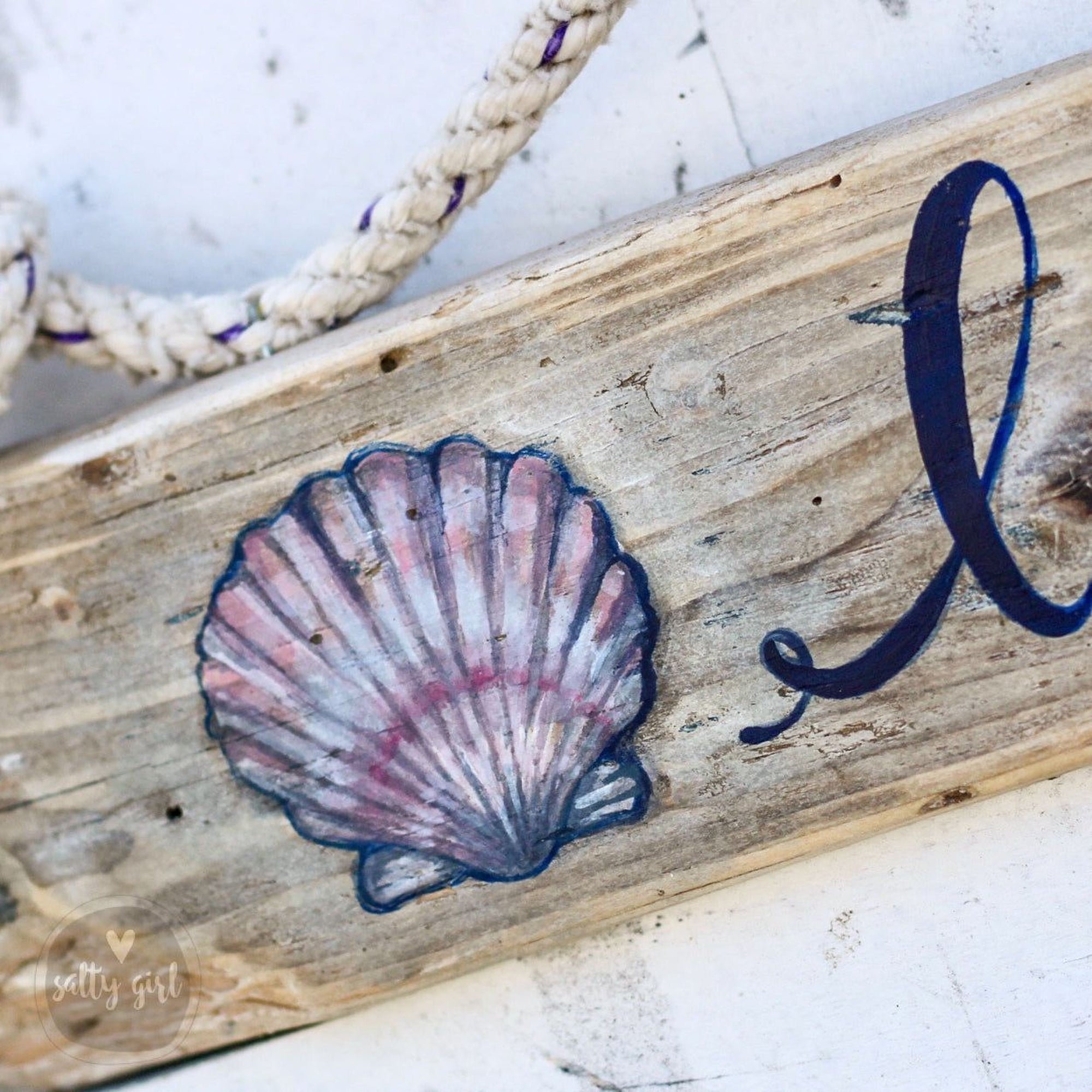 A wooden sign with a painted seashell and a blue cursive letter "L" on it.