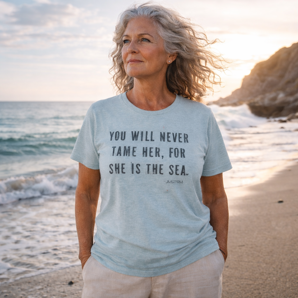 A woman with long, gray hair stands on a beach, wearing a light blue t-shirt with the text "You will never tame her, for she is the sea" printed on it.