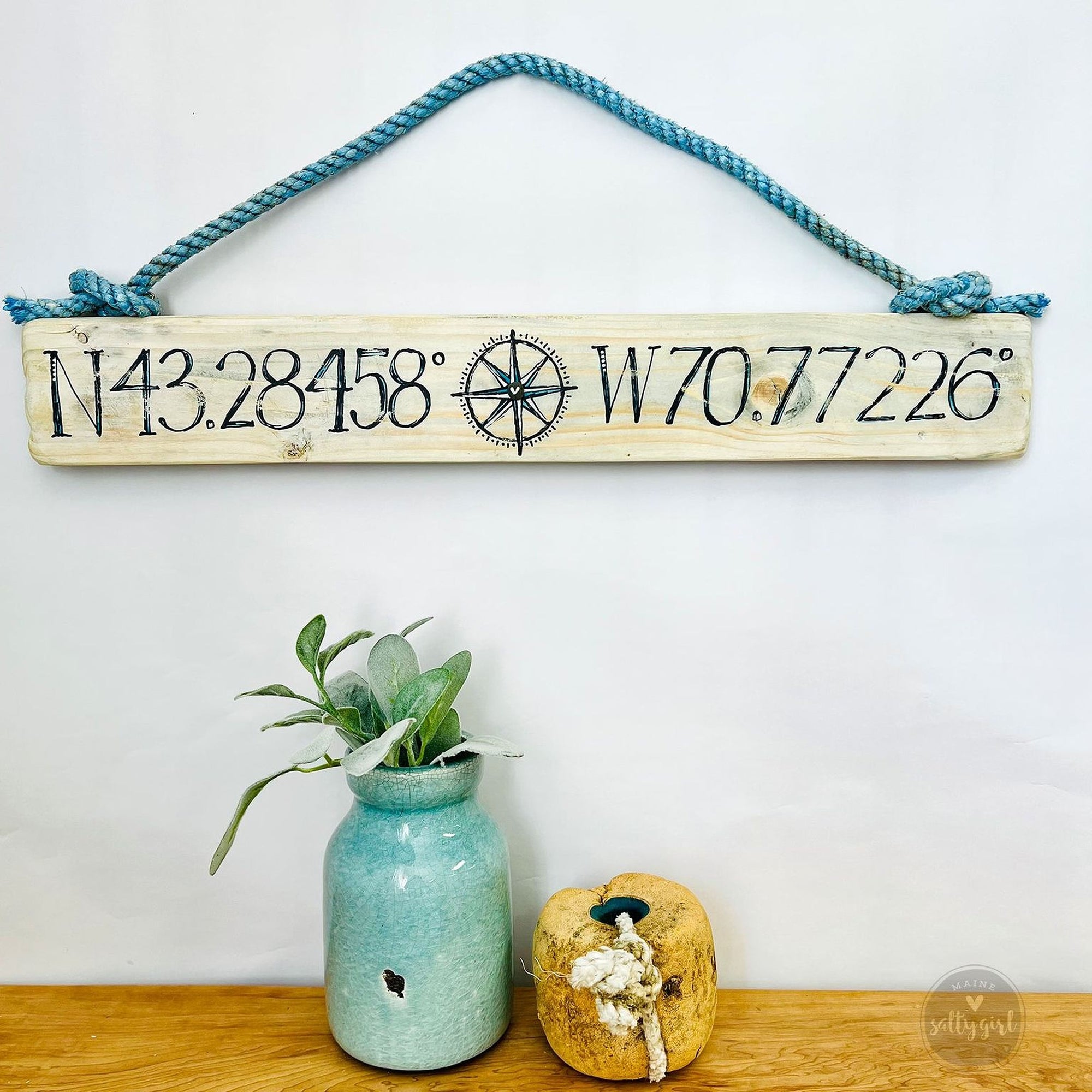 a wooden sign with a compass rose and the numbers "N43.284558 W70.77226" hanging on a wall, accompanied by a small blue vase with a plant and a small wooden object on a shelf.