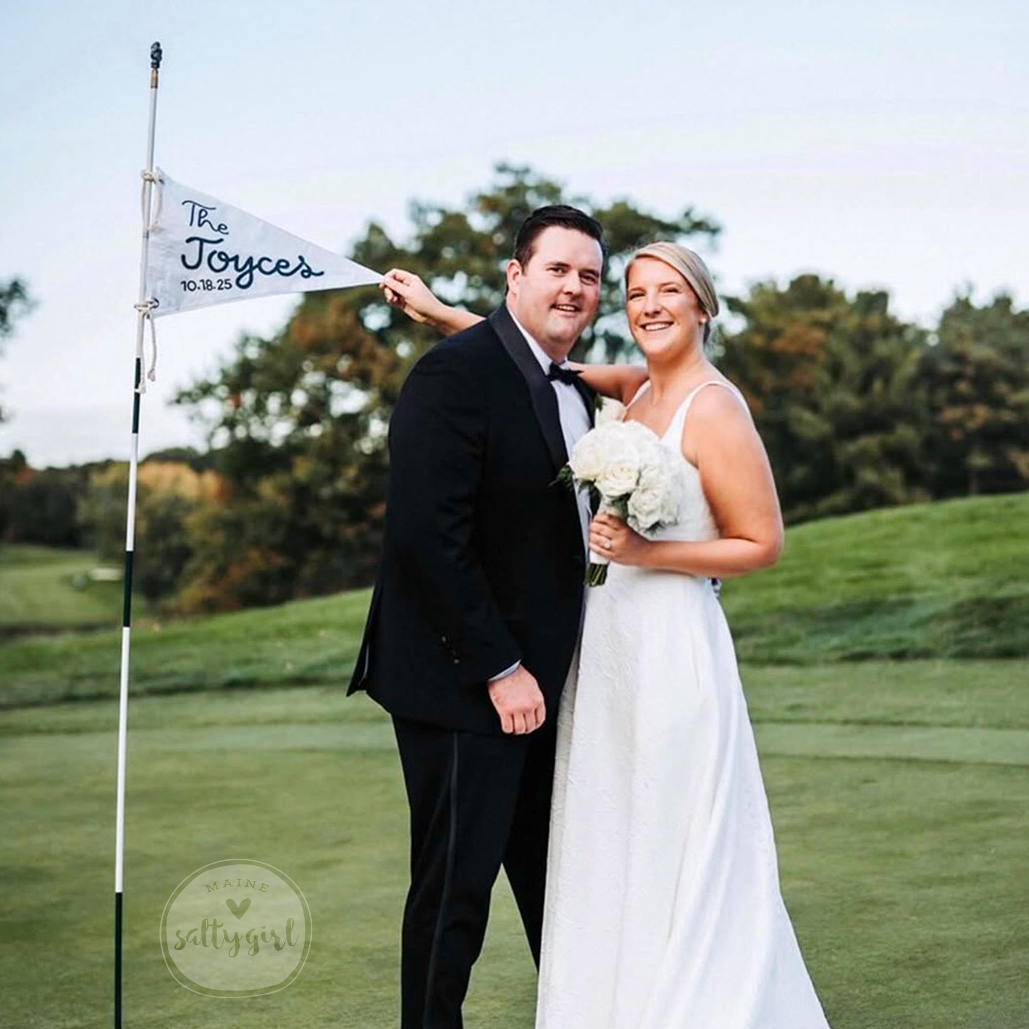 A newlywed couple poses for a photo on a golf course, with the bride holding a bouquet of white flowers and the groom wearing a tuxedo.