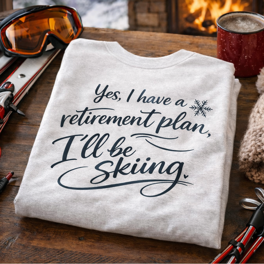 A white t-shirt with a humorous retirement plan and skiing message, along with ski gear and a mug, is displayed on a wooden surface.
