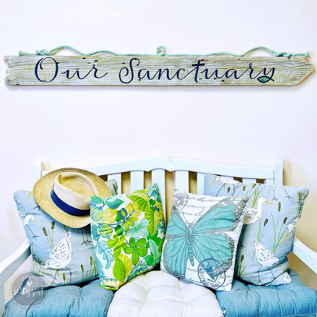 a wooden sign with the text "Our Sanctuary" hanging on a wall above a white bench with colorful pillows and a straw hat.
