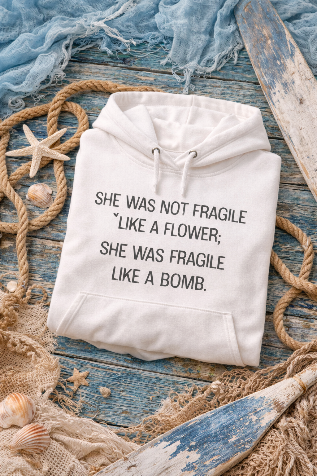A white hoodie with a humorous text printed on it, lying on a wooden surface surrounded by seashells, rope, and a starfish.