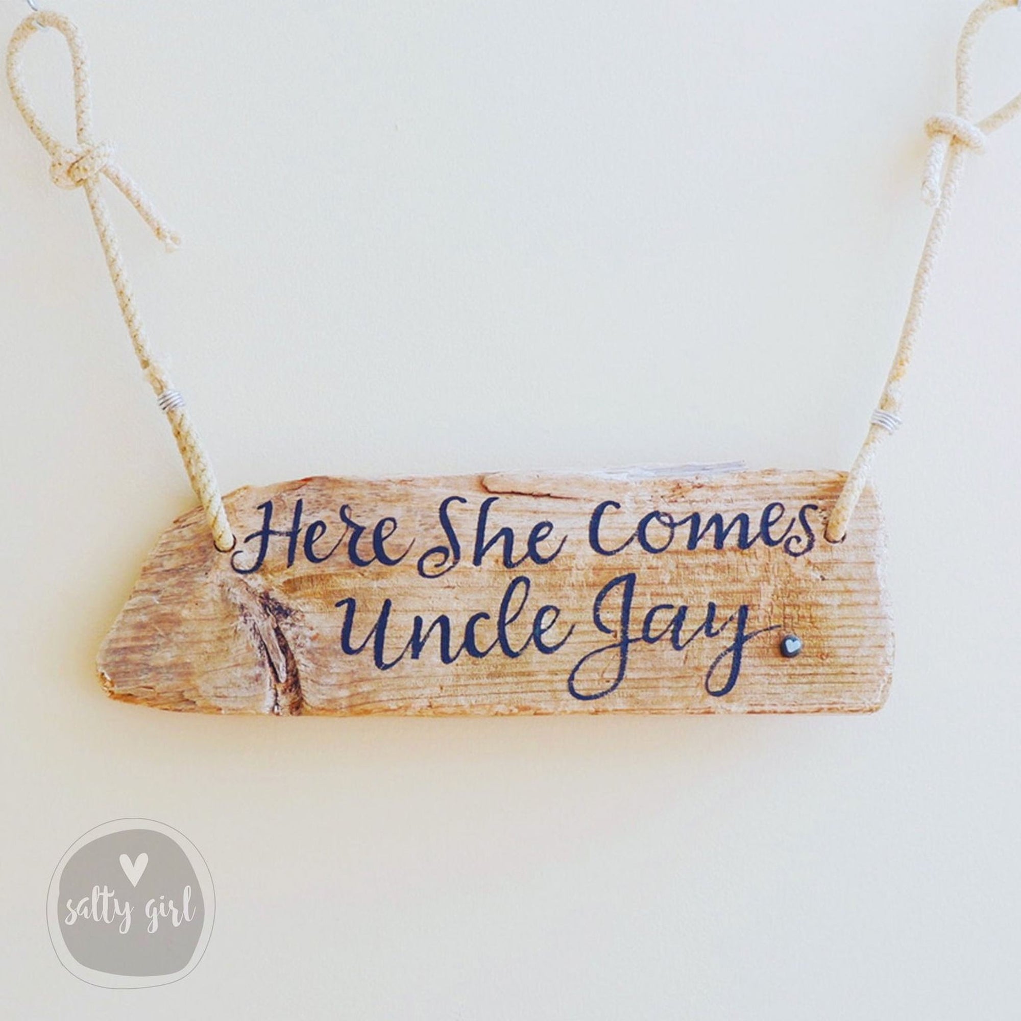 A wooden sign with the text "Here She Comes Uncle Jay" written on it, hanging from a rope.