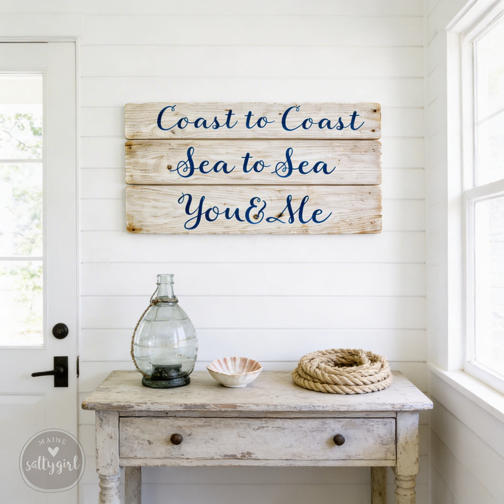 a rustic wooden sign with the phrase "Coast to Coast Sea to Sea You & Me" hanging on a white wall above a small table with various items on it, including a glass vase, a bowl, and a rope.