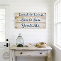 a rustic wooden sign with the phrase "Coast to Coast Sea to Sea You & Me" hanging on a white wall above a small table with various items on it, including a glass vase, a bowl, and a rope.