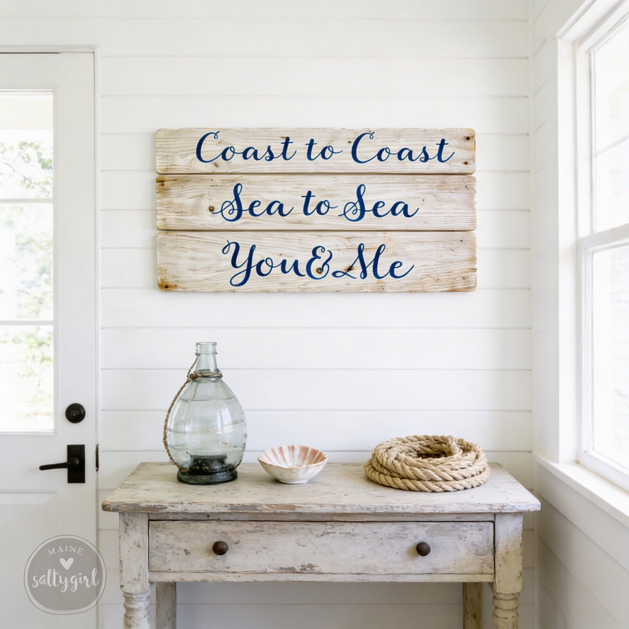 a rustic wooden sign with the phrase "Coast to Coast Sea to Sea You & Me" hanging on a white wall above a small table with various items on it, including a glass vase, a bowl, and a rope.