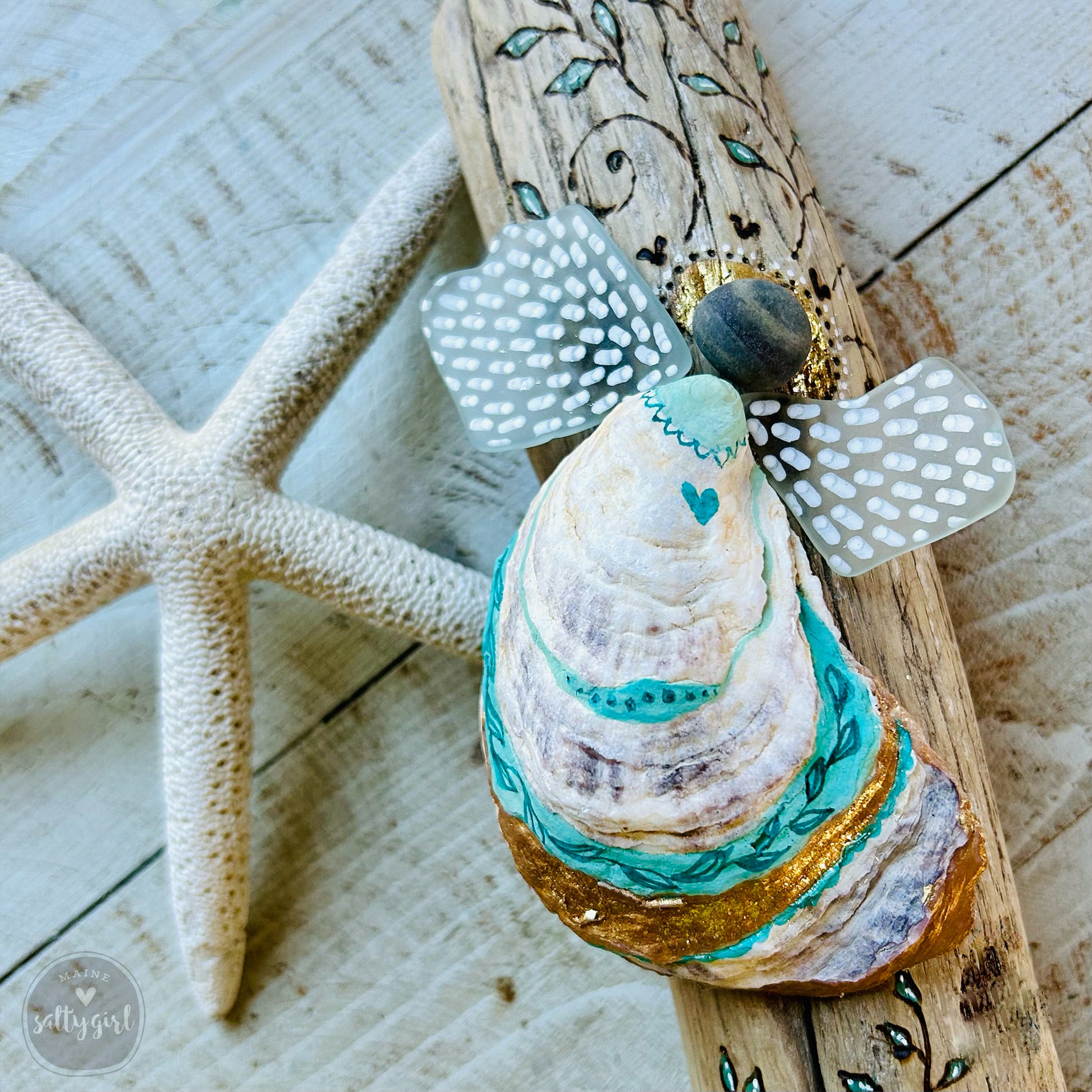 A handcrafted wooden angel figure made from a seashell, adorned with a heart and decorated with a floral pattern, is displayed on a wooden surface alongside a starfish.