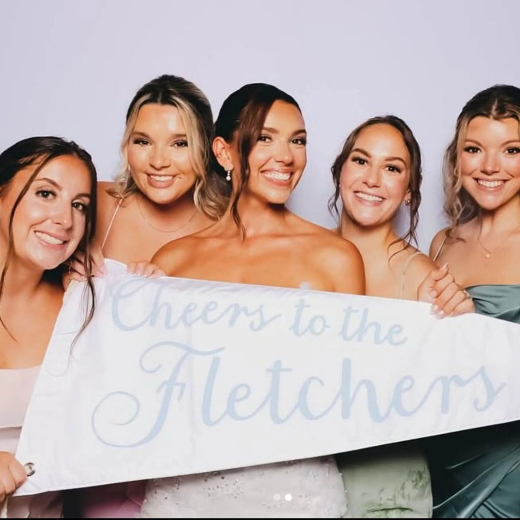 Five women holding a wedding pennant that reads 'Cheers to the Fletchers' against a plain background
