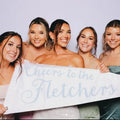 Five women holding a wedding pennant that reads 'Cheers to the Fletchers' against a plain background