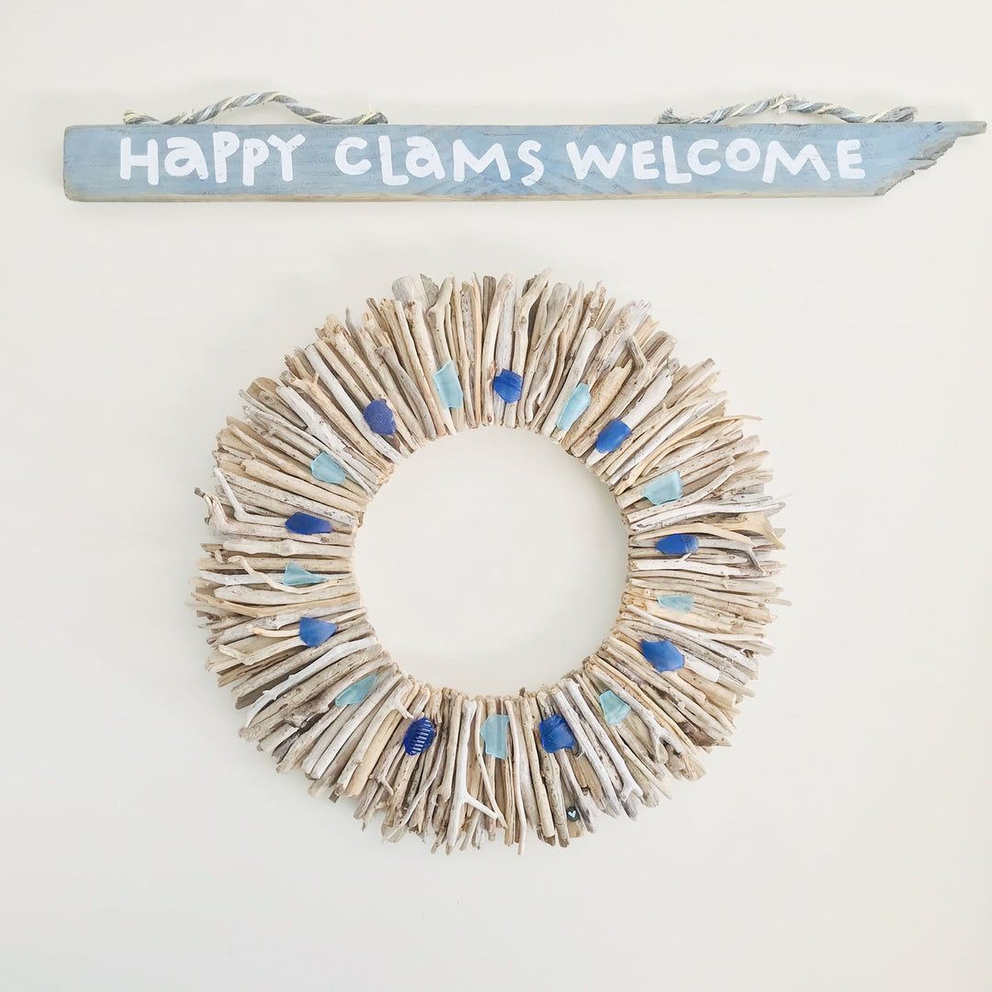 A wreath made of driftwood and blue stones, hanging on a wall with a sign that says "Happy Clams Welcome".