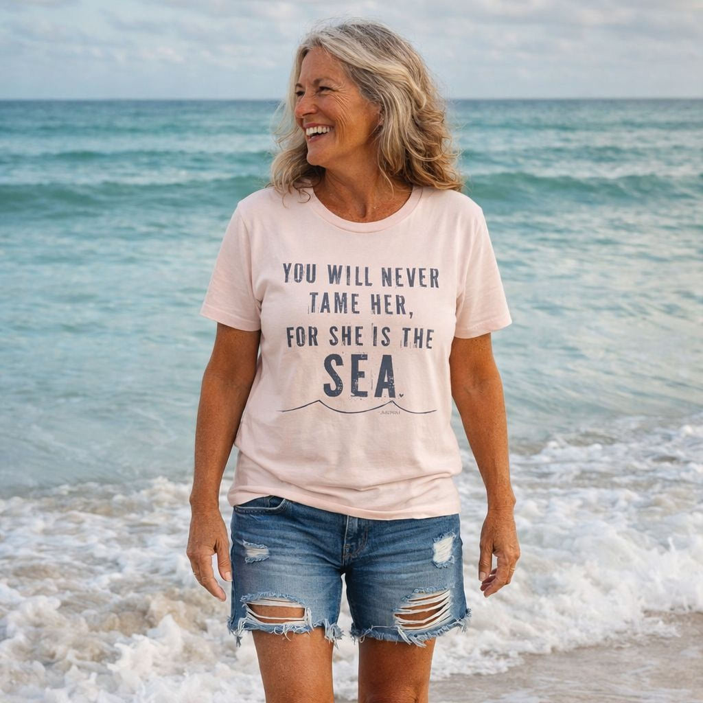 A woman standing on a beach, smiling and wearing a t-shirt with text and denim shorts, with the ocean in the background.