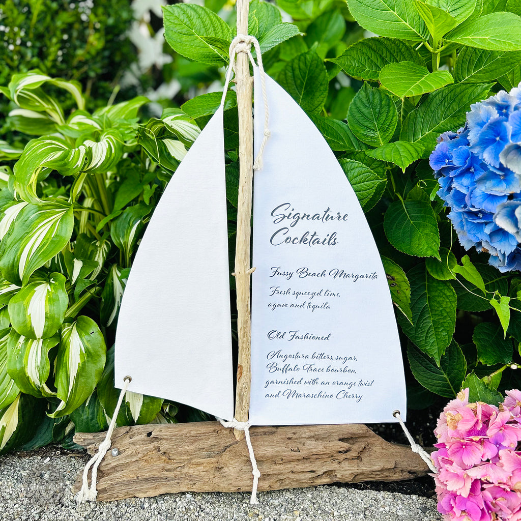A white sailboat-shaped sign with black text is displayed in a garden setting, surrounded by lush greenery and colorful flowers.