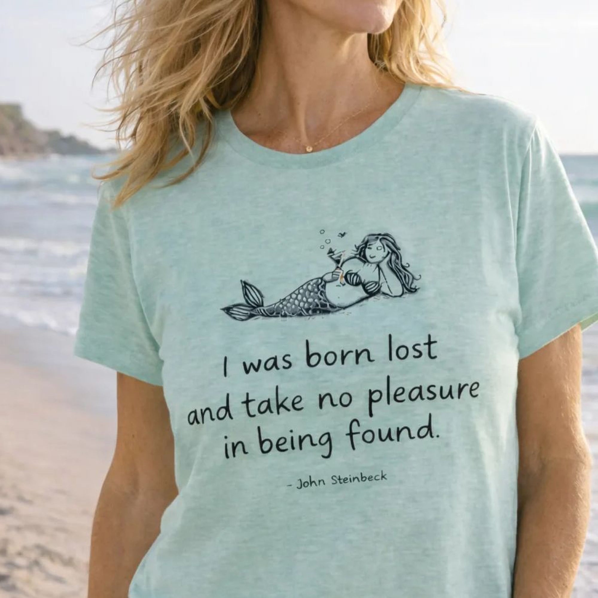 A woman wearing a light blue Maine Salty Girl t-shirt with a mermaid graphic and a quote printed on it, standing on a beach.