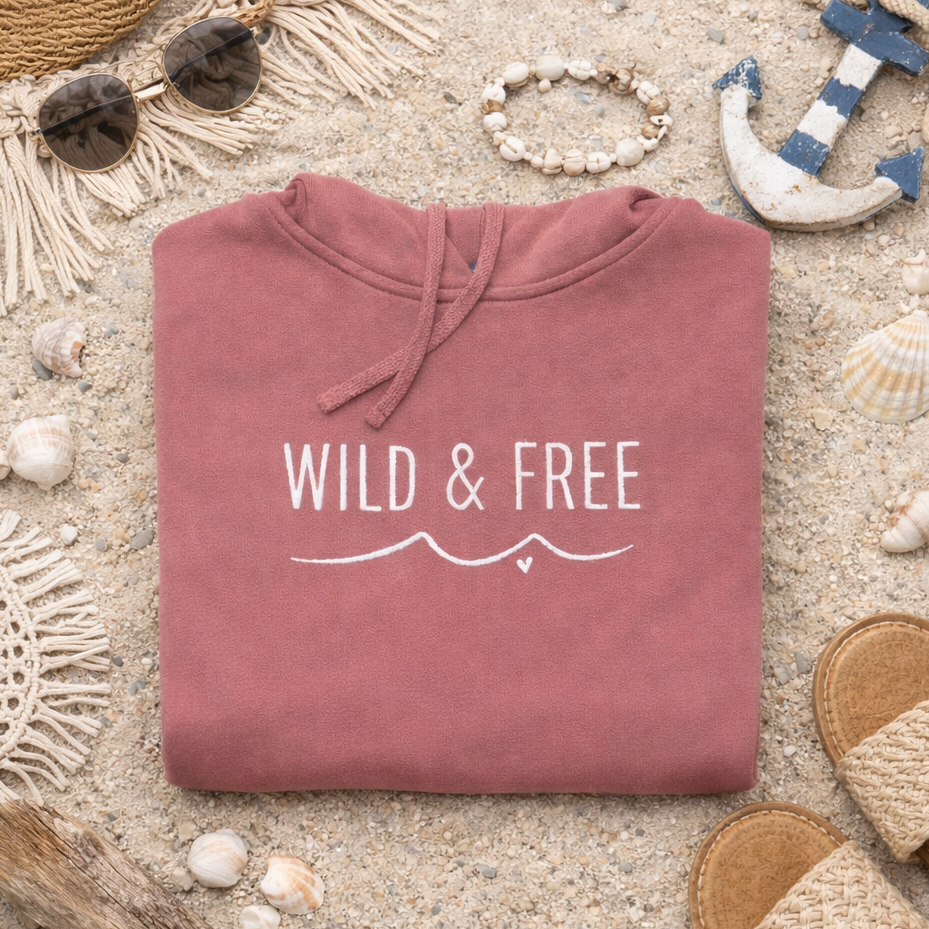 A pink hoodie with the text "WILD & FREE" printed on it, along with a pair of sandals, sunglasses, and a necklace, all laid out on a sandy beach.