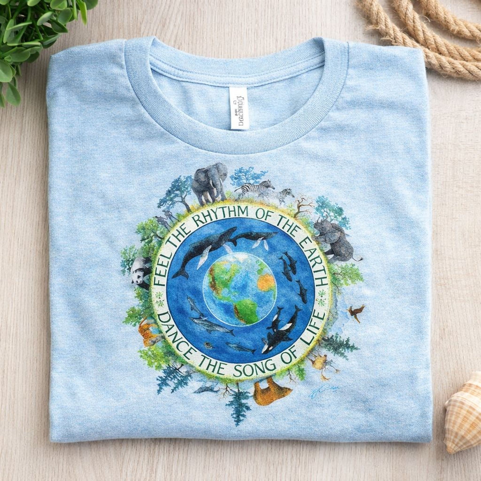 A light blue Maine Salty Girl t-shirt with a circular design featuring a depiction of the Earth surrounded by various animals and plants, along with the text "Feel the rhythm of the Earth, dance the song of life".