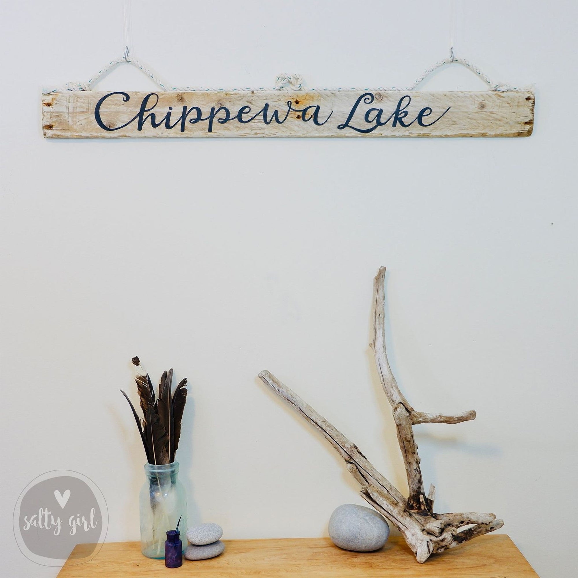 a wooden sign that reads "Chippewa Lake" hanging on a wall, along with a wooden table displaying various items such as a vase with feathers, a rock, and a branch.