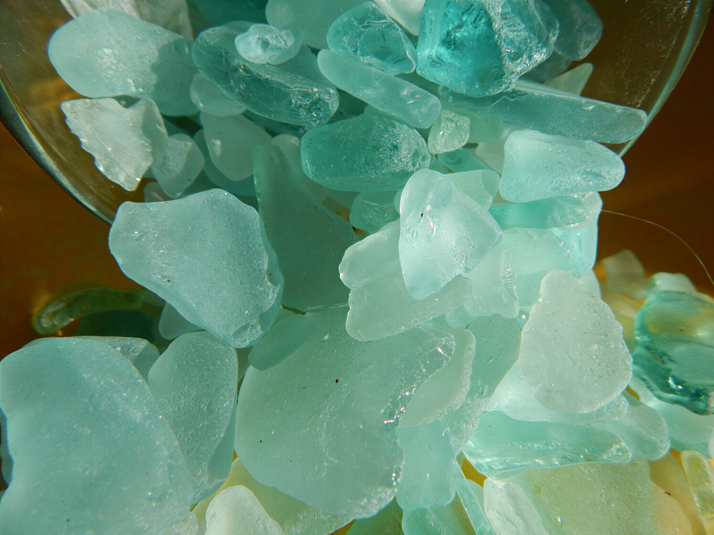 A close-up image of a pile of various shades of blue and green sea glass, with some pieces appearing to be in motion as they are being poured or dropped into a container.