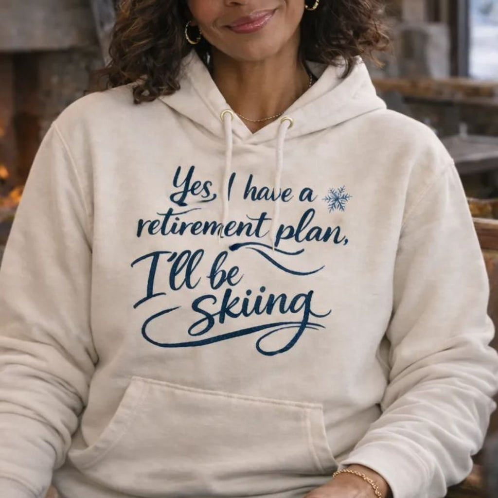 A woman wearing a white Maine Salty Girl hoodie with a humorous text and a pair of ski goggles, smiling at the camera in a cozy cabin setting.