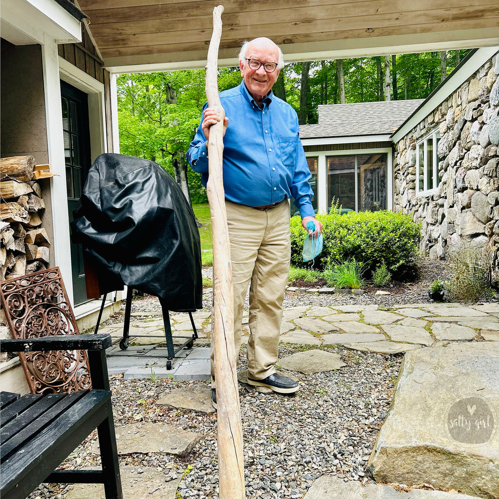 An elderly man stands in a backyard, holding a large wooden stick. He is wearing a blue shirt and khaki pants. The backyard has a stone patio, a grill, and a wooden bench.