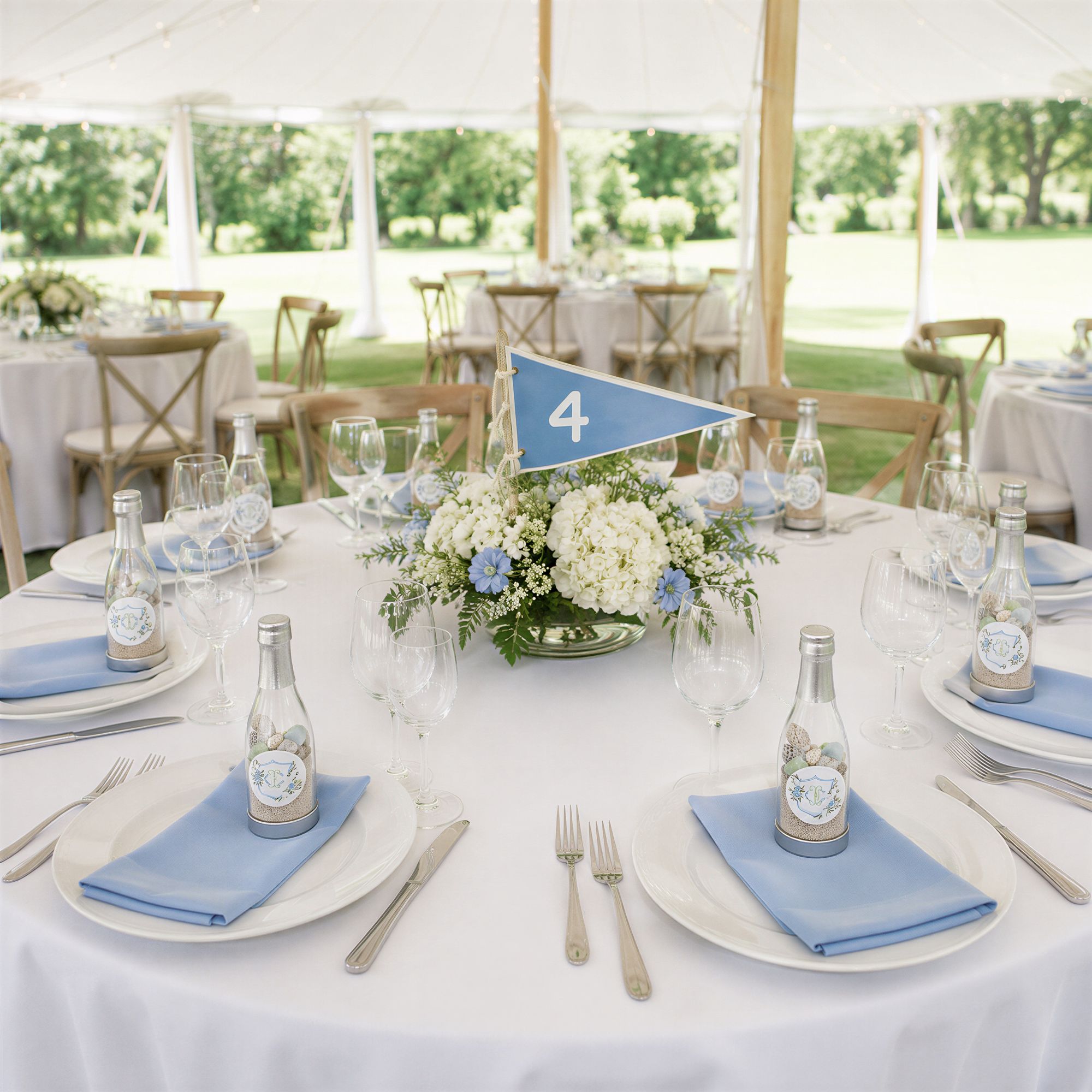 a beautifully set dining table with place settings, including plates, silverware, and glassware, arranged under a white tent. The table is adorned with a floral centerpiece and a blue flag, creating a festive atmosphere.