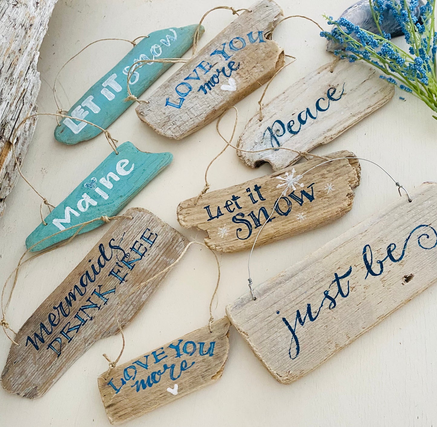 a collection of handcrafted wooden signs with various sayings and designs, including "Let it snow", "Love you more", "Just be", and "Maine". The signs are hanging on a surface, creating a visually appealing and decorative arrangement.